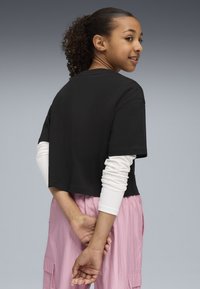 Black cropped t-shirt made of cotton, paired with a long white sleeve underneath, worn with pink cargo pants. Smooth texture, casual fit.