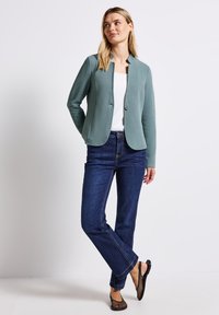 Žena v texturovaném teal blejzru přes bílý top, tmavě modrých džínách a černých síťovaných slip-on botách stojící proti bílému pozadí.