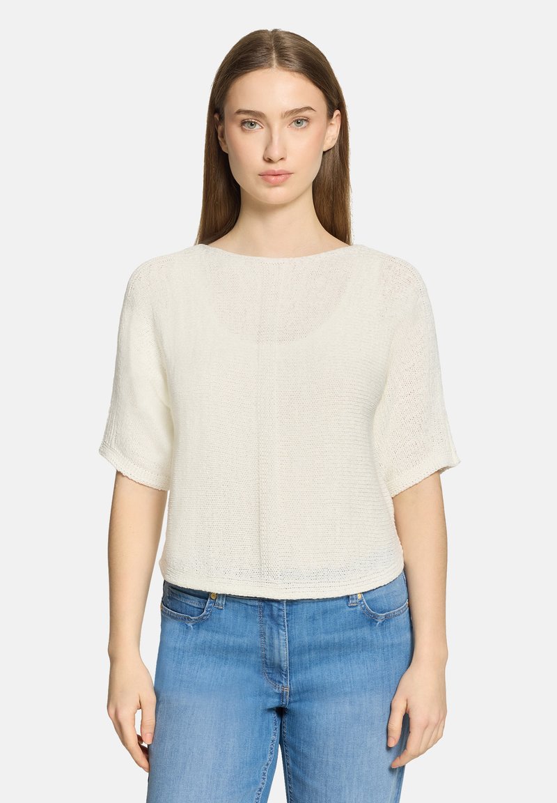 Junge Frau mit glatten braunen Haaren, die ein weißes kurzärmeliges Strickoberteil und blaue Jeans trägt, vor einem schlichten Hintergrund stehend.
