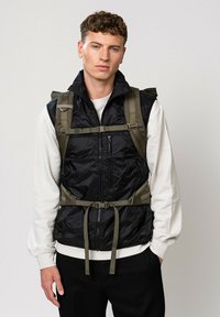 Gilet senza maniche nero con texture lucida, zip frontale, spalline e cintura regolabili verde oliva, indossato sopra una camicia a maniche lunghe bianca.