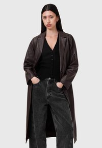 Cappotto trench in pelle marrone con revers a punta, maniche lunghe e apertura frontale. Indossato sopra una maglietta nera a coste e jeans grigi a vita alta.