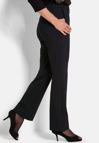 Vrouw in zwarte, strakke broek en zwarte, open teen hoge hakken, staand met één been iets naar voren.