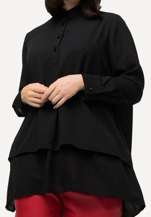 Kobieta w nałożonej czarnej, zapinanej na guziki koszuli z długim rękawem oraz czerwonych skórzanych spodniach, ręce splecione przy talii.