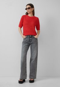 Rode gebreide top met korte mouwen, gecombineerd met hooggewaist grijze wijde jeans en zwarte plateau-sandalen. Zonnebril maakt de look compleet.