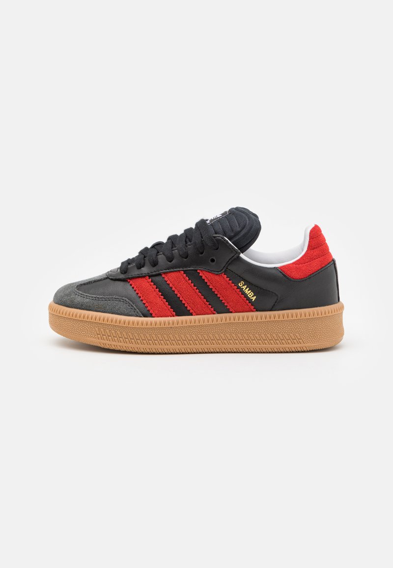 Svarte sneakers med røde striper, teksturert semsket skinn og lær overdel, gummisåle og polstret tunge. Har logo-detaljer på siden.
