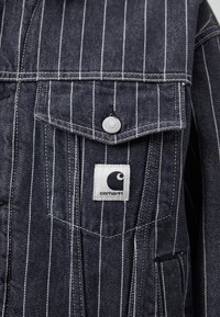 Donker denimjasje met witte verticale strepen, voorzien van een borstzak, metalen drukknoop en een vierkante logopatch aan de linkerkant.