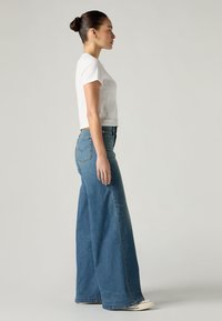 Λευκό κοντομάνικο cropped t-shirt, συνδυασμένο με φαρδιά μπλε τζιν παντελόνια. Τα τζιν διαθέτουν επένδυση τσέπης και ίσια, φαρδιά εφαρμογή.