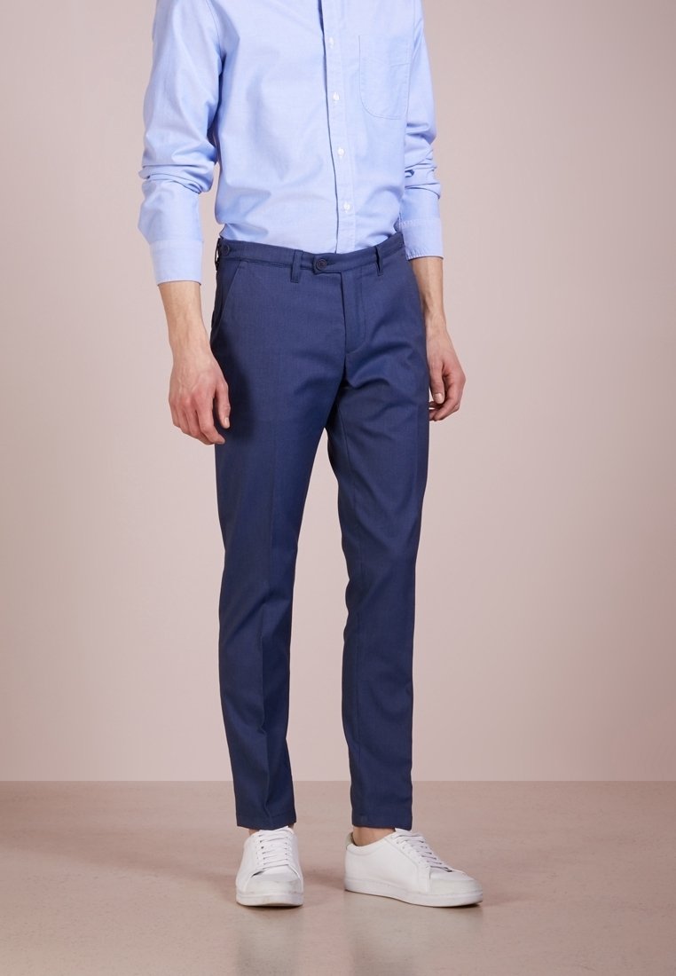 Mężczyzna ubrany w jasnoniebieską koszulę z guzikami, granatowe slim-fit spodnie i białe sneakersy, stojący na beżowej podłodze.