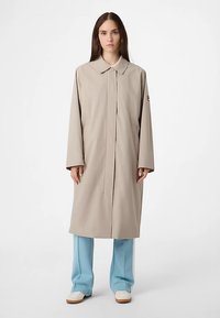 Lysebeige lang frakke med krave, lige snit og lukning med knapper. Den har lange ærmer og sidelommer.