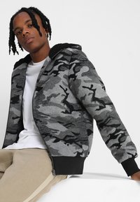 Jachetă cu glugă, cu model de camuflaj gri și accente negre, cu un material moale. Purtați peste un tricou alb, asortată cu pantaloni bej.