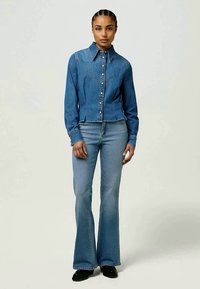 Farmerdzseki galléros nyakkal és gombos elülső résszel, amit harangszárú farmernadrággal párosítanak; mindkettő közepes kék mosással, finom kopásokkal.