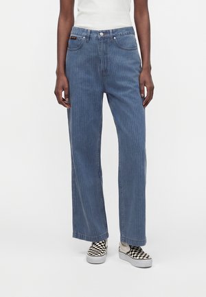 Person iført højtaljede, løstsiddende blå jeans med subtile vertikale striber, hvid top og sort-hvide slip-on sko med ternet mønster.