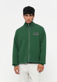 Groene zip-up jas van een zacht materiaal, met een hoge kraag en een rechthoekig logo-patch op de linkerkant van de borst.