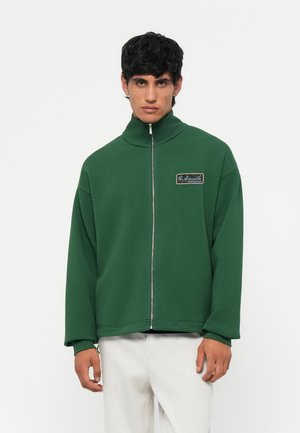 Green zip-up jacket made of a soft material, featuring a high collar and a rectangular logo patch on the left chest.