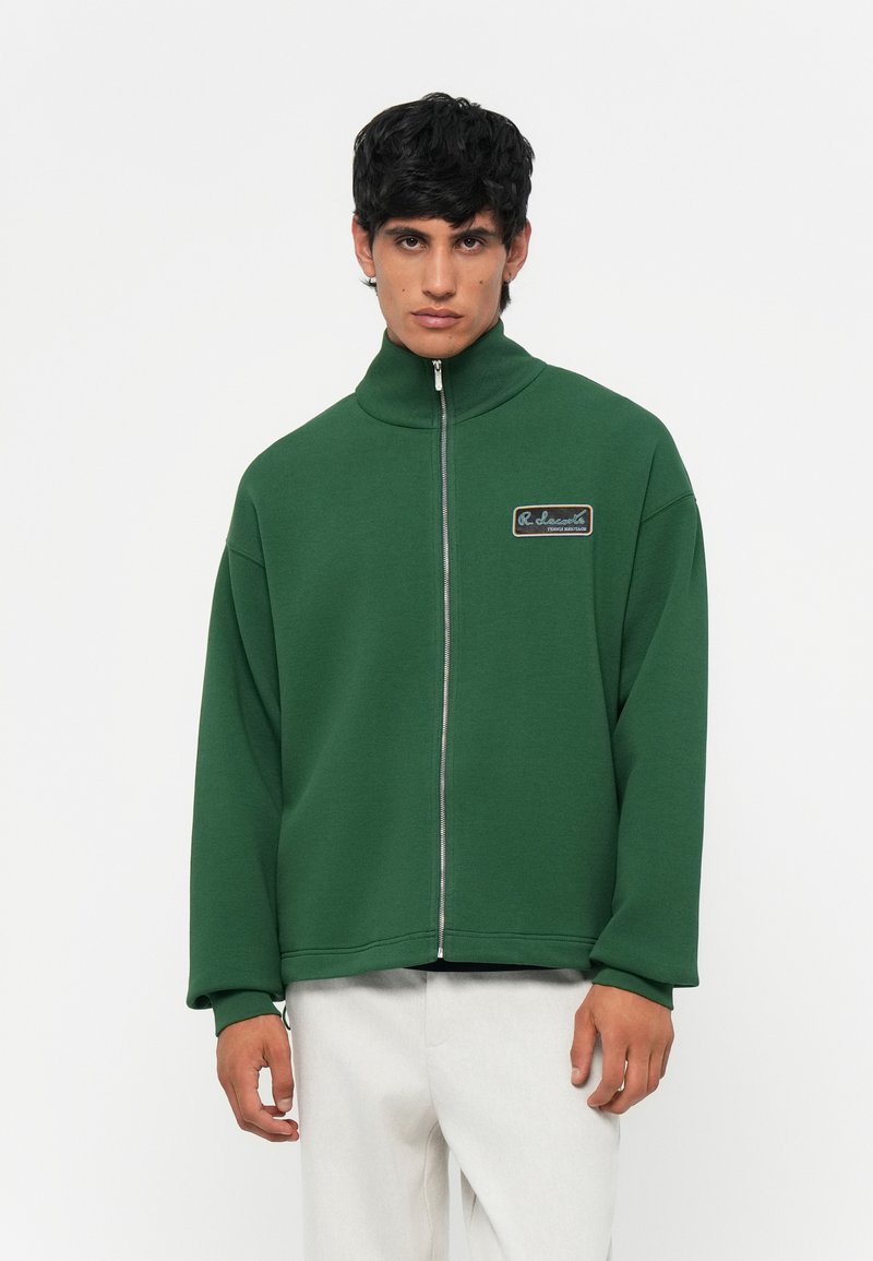 Groene zip-up jas van een zacht materiaal, met een hoge kraag en een rechthoekig logo-patch op de linkerkant van de borst.