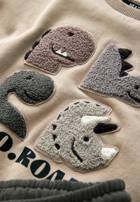 Felpa beige con quattro toppe di dinosauro testurizzate in marrone, grigio e crema, con un dettaglio testuale stampato sotto. Tessuto morbido.