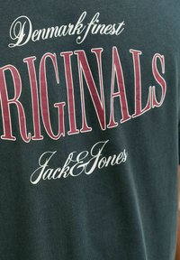 Camiseta de algodón verde oscuro con texto blanco y burdeos: "Los mejores ORIGINALS de Dinamarca Jack & Jones" en diferentes tipografías y tamaños.