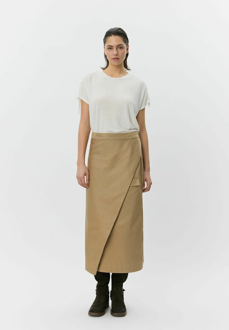 Femme aux cheveux lissés vers l'arrière portant un haut blanc à manches courtes, une jupe portefeuille beige, des bottes noires, debout devant un fond blanc uni.
