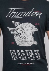 T-shirt noir avec un graphic usé d'un casque ailé. Le texte indique "Thunder," "Race," et "Born to be Free," en blanc et rouge. Fabriqué aux États-Unis.