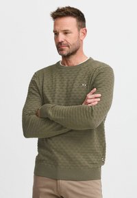 Olijfgroene trui met een diamanttextuur, geribde manchetten en zoom, en een klein, multikleurig logo op de borst.
