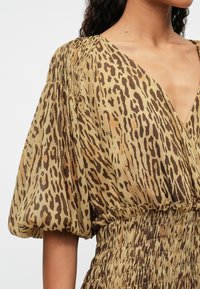 Šaty s leopardím potiskem, které mají hluboký V-výstřih, nabírané rukávy a nařasený pas. Vyrobeny z lehkého materiálu s hladkou texturou v přírodních barvách.