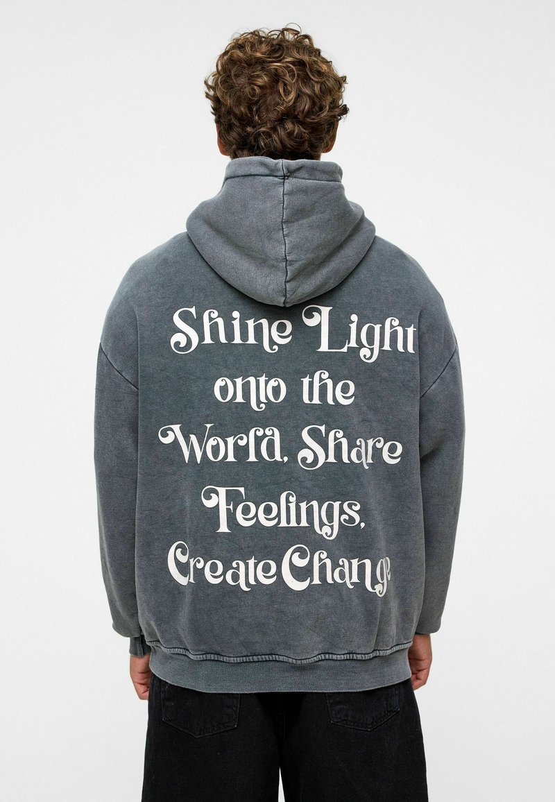 Grijze oversized hoodie met een voorzak in kangoeroestijl, voorzien van witte gedrukt tekst op de achterkant die inspirerende zinnen bevat.