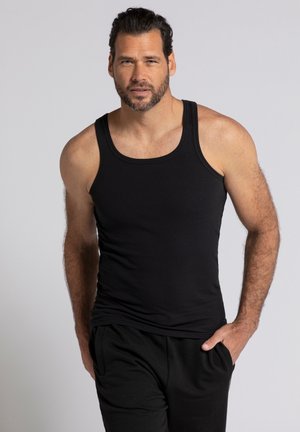 Schwarzes geripptes Tanktop mit breiten Trägern, figurbetonter Form und weichem Stoff, getragen mit schwarzen Hosen, Modell steht mit der Hand in der Tasche.