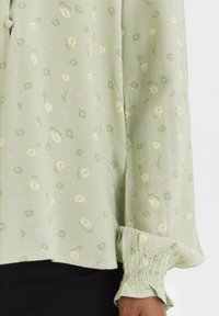 Lichtgroene blouse met kleine bloemenpatronen in geel en paars, met een knoopdetail en gerimpelde manchetten voor textuur.