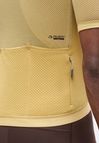 Gros plan d’un maillot de cyclisme en tissu texturé jaune moutarde avec une petite poche zippée et le logo Polartec Delta dans le dos.