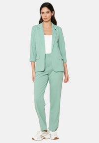 Mėtų žalia sakai ir atitinkančios kelnės, turinčios laisvą siluetą, sukamuosius rankovėmis ir kišenėmis. Stiliuota su balta viršutine dalimi ir kasdai sportbačiais.