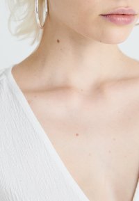 Bijela teksturirana majica s dubokim dekolteom, srebrne naušnice u obliku obruča i vidljiva koža, koja pokazuje lagane pjegice i prirodne detalje tena.