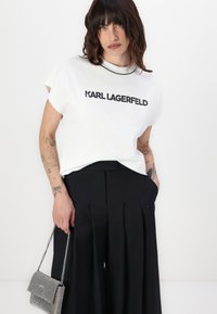 Baltas kokvilnas T-krekls ar melnu "KARL LAGERFELD" logotipu, īsām piedurknēm. Saskaņots ar augsto vidukli melnām veļas biksēm un sudraba ķēdes somu.