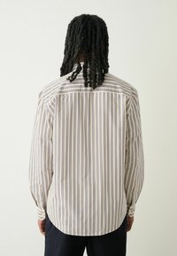 Pruhovaná košile s dlouhým rukávem v béžové a námořnické barvě, s volným střihem. Má zapínání na knoflíky a zaoblený lem, vyrobeno z lehké tkaniny.