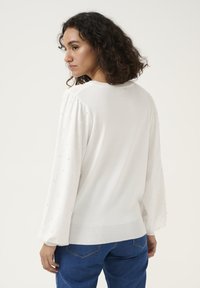 Witte trui met opbollende mouwen, voorzien van een gestructureerd patroon en kleine decoratieve kraaltjes, gecombineerd met een blauwe spijkerbroek, weergegeven vanuit de achterkant.