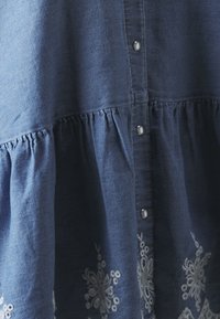Blå denimkjole med innsnørt liv, sølvknapper og hvit blomsterbroderi nederst. Myk tekstur og avslappet passform.