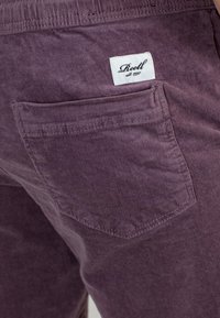 Pantalones de pana morados con un bolsillo trasero y una etiqueta de marca blanca con texto negro. La textura es acanalada con líneas verticales.