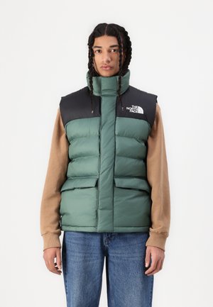 Green and black puffer vest with high collar, two front pockets, and logo on the left chest. Worn over a brown long-sleeve garment.