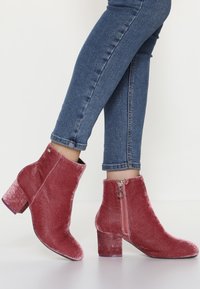 Piernas en jeans skinny azules que llevan botas de terciopelo rosa polvo hasta el tobillo con tacones de bloque y cremalleras laterales, de pie sobre un suelo blanco.