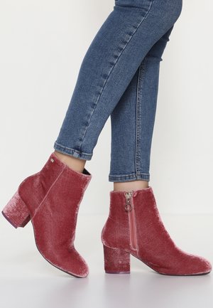 Jambes dans un jean skinny bleu portant des bottines en velours rose poussiéreux avec des talons carrés et des fermetures éclair sur le côté, debout sur un sol blanc.