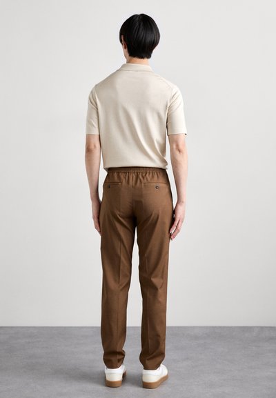 Lys beige kortærmet polo-shirt parret med brune elastiske bukser og hvide sneakers, vist bagfra mod en neutral baggrund.