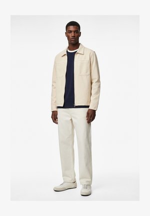 Mężczyzna w beżowej kurtce, granatowej koszuli, kremowych spodniach i białych trampkach, stojący na tle gładkiego białego tła.