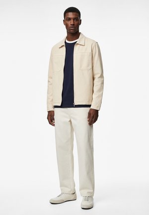 Mann trägt eine beige Jacke, ein marineblaues Hemd, cremefarbene Hosen und weiße Turnschuhe, steht vor einem schlichten weißen Hintergrund.