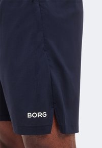 Σκούρο μπλε αθλητική βερμούδα με πλευρική σχισμή και λευκό λογότυπο "BORG", φορεμένη από ένα άτομο, που δείχνει την υφή του υφάσματος και το δέρμα του ποδιού κάτω από το στρίφωμα.