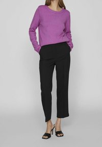 Lila tröja med ribbad rund halslinning, kombinerad med svarta åtsittande byxor. Modellen har på sig svarta öppna klackskor och har h änderna i fickorna.