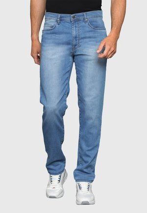 Uomo che indossa jeans blu scoloriti e sneaker bianche, in piedi con le mani rilassate lungo i fianchi su uno sfondo chiaro e semplice.