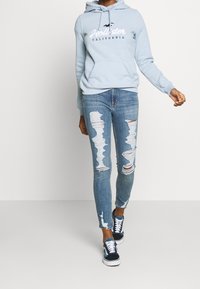 Felpa con cappuccio azzurro chiaro con tasca frontale e logo, jeans blu sbiaditi con molteplici strappi e scarpe da ginnastica nere con dettagli bianchi.