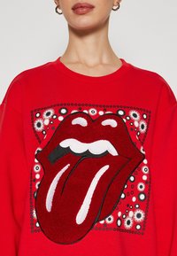 Sweat-shirt rouge avec un logo brodé proéminent représentant des lèvres ouvertes avec une langue, entouré d'accents aux motifs en noir et blanc.