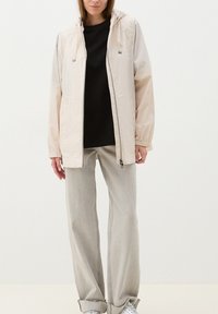 Chaqueta ligera beige con capucha, puños elásticos y cierre frontal con cremallera, llevada sobre una camisa negra y combinada con pantalones de pierna ancha grises.