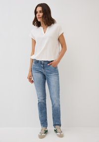 Kvinna med axellångt brunt hår, klädd i en vit kortärmad blus, blå jeans och beige sneakers med gröna ränder, stående med handen i fickan.