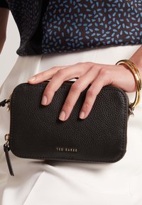 Schwarze Clutch aus strukturiertem Leder mit goldfarbenen Beschlägen und Logo. Verfügt über einen Reißverschluss und eine kleine Quastenverzierung. Wird in der Hand gehalten.